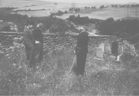 A black and white photo of three men, two in uniform and one standing with a crutch, stand in a field with gravestones, fields and trees beyond them in landscape.